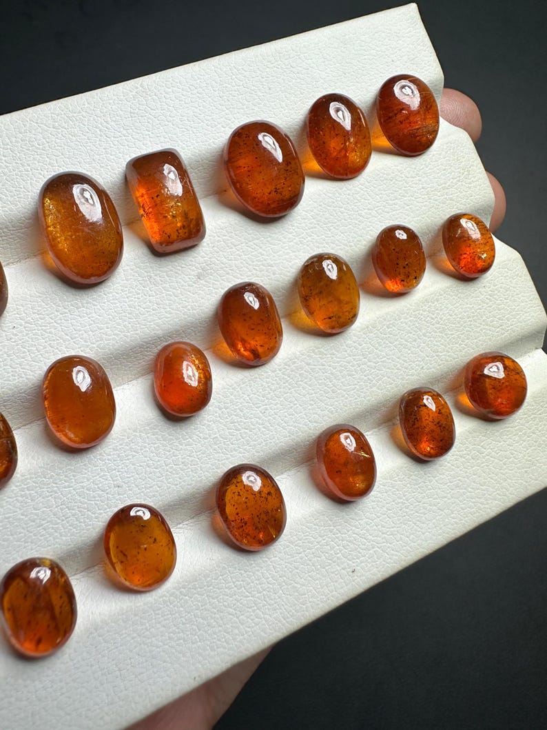 May include: A collection of 15 oval and rectangular shaped gemstones in various shades of brown. The gemstones are displayed on a white surface.