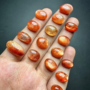May include: A collection of 17 oval-shaped sunstone cabochons in various shades of orange and red. The stones are translucent and have a shimmering, iridescent effect.