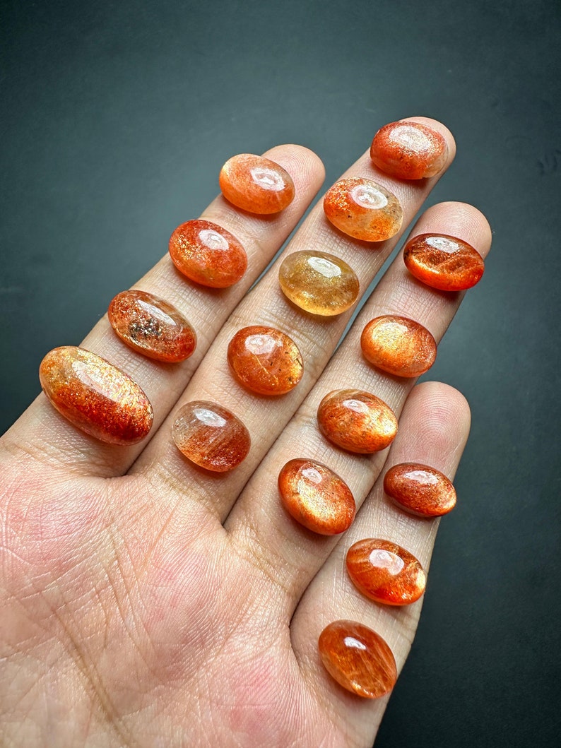 May include: A collection of 17 oval-shaped sunstone cabochons in various shades of orange and red. The stones are translucent and have a shimmering, iridescent effect.