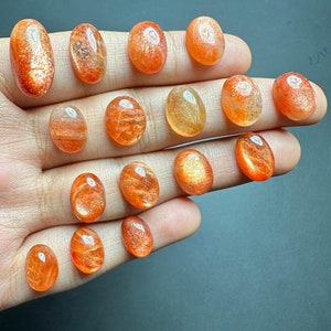 May include: A collection of 15 oval-shaped sunstone gemstones in various shades of orange and yellow. The stones are smooth and polished, with a shimmering, iridescent effect.