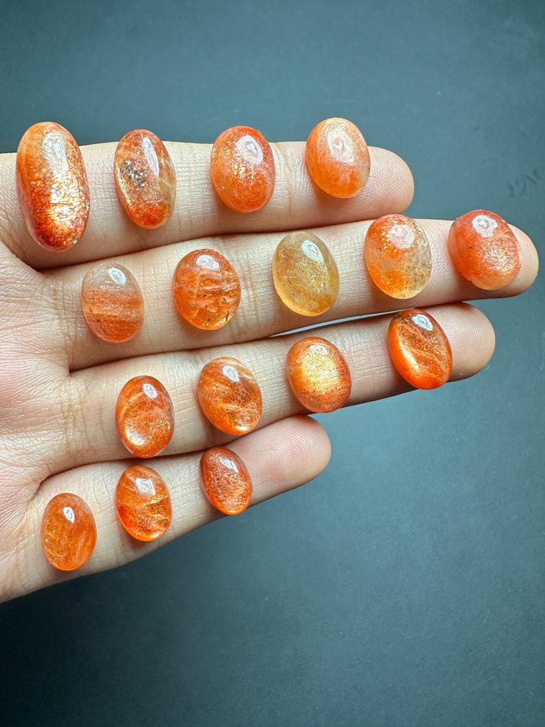 May include: A collection of 15 oval-shaped sunstone gemstones in various shades of orange and yellow. The stones are smooth and polished, with a shimmering, iridescent effect.