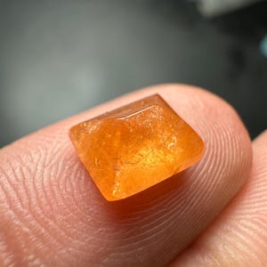 May include: A square-cut, orange gemstone with a faceted top. The gem is held between two fingers, showcasing its color and shape. The gemstone appears to be translucent, with a slightly rough texture.
