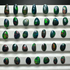 May include: A collection of 36 loose black opal gemstones, each with a unique green and red iridescent sheen. The opals are arranged in rows on a white surface.