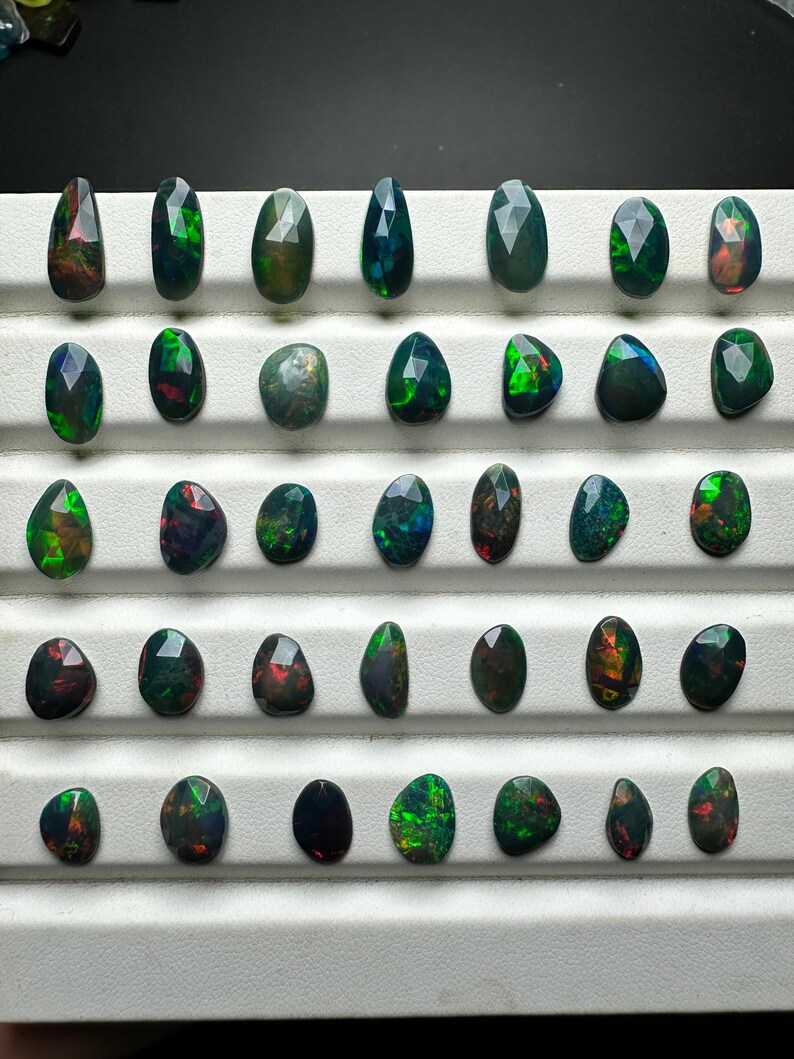 May include: A collection of 36 loose black opal gemstones, each with a unique green and red iridescent sheen. The opals are arranged in rows on a white surface.