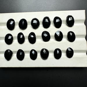 May include: A collection of 16 black gemstones, likely obsidian, displayed on a white tray. The gemstones are faceted and have a smooth, polished finish.
