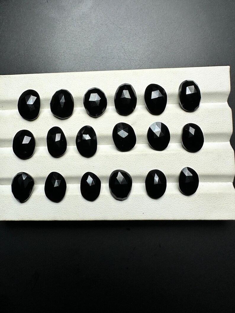 May include: A collection of 16 black gemstones, likely obsidian, displayed on a white tray. The gemstones are faceted and have a smooth, polished finish.
