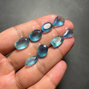 May include: Eight faceted, light blue gemstones of varying shapes and sizes are displayed. The gemstones are held in a hand against a dark background. The gems include oval, pear, and teardrop shapes, each with a unique cut and sparkle.