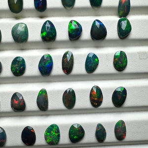 May include: A collection of 25 black opal gemstones, each with a unique green, red, and blue iridescence. The opals are displayed on a white surface.