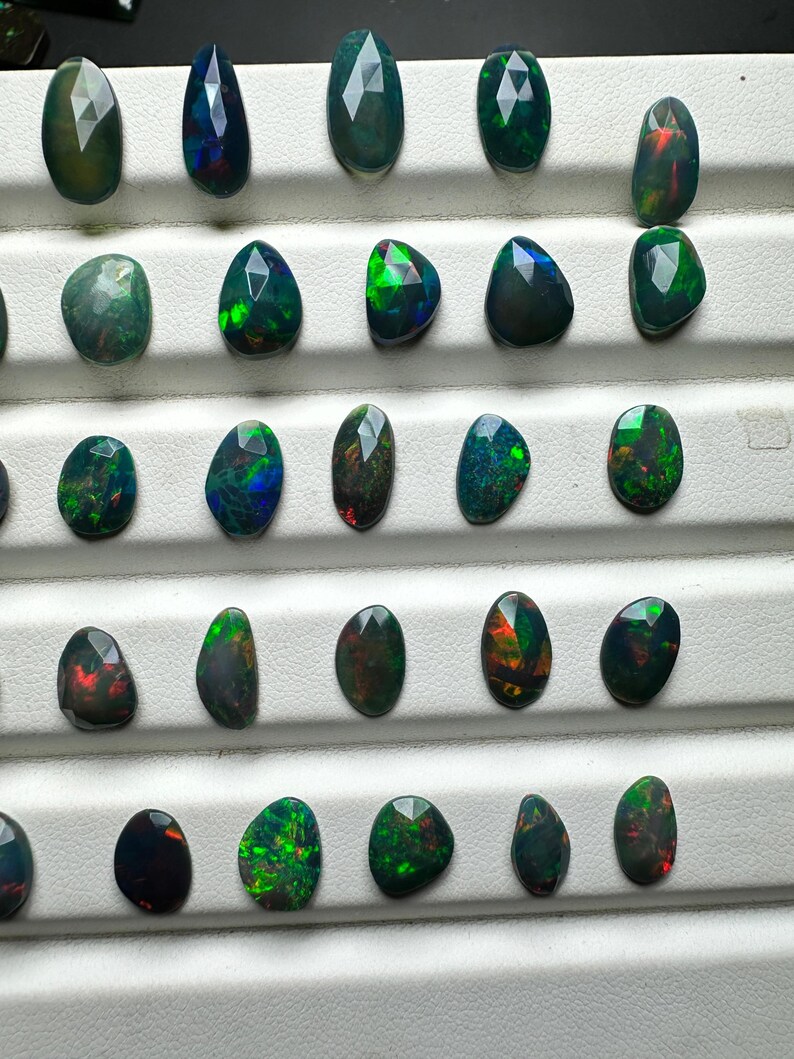 May include: A collection of 25 black opal gemstones, each with a unique green, red, and blue iridescence. The opals are displayed on a white surface.
