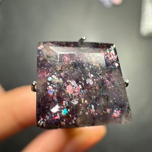 May include: A square-shaped ring featuring a dark purple, translucent stone with embedded iridescent glitter particles that reflect pink, blue, and silver light. The stone is held in place by a simple silver-toned metal prong setting.