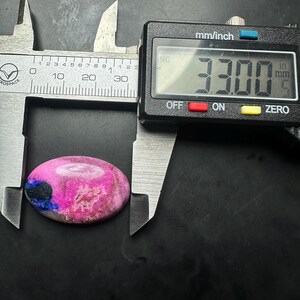 May include: An oval-shaped cabochon stone featuring vibrant pink and magenta hues with a distinct dark blue inclusion on the left side. A digital caliper is positioned over the stone, displaying a measurement of 33.00 millimeters in length.