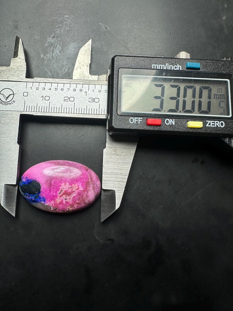 May include: An oval-shaped cabochon stone featuring vibrant pink and magenta hues with a distinct dark blue inclusion on the left side. A digital caliper is positioned over the stone, displaying a measurement of 33.00 millimeters in length.