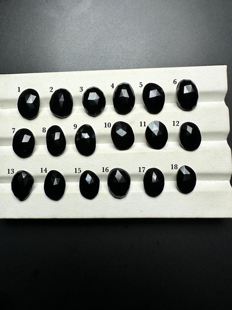 May include: A set of 18 black gemstones, numbered 1 through 18, arranged in rows on a white surface. The gemstones are faceted and have a shiny, polished finish.