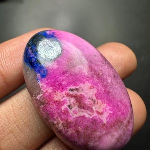 May include: An oval-shaped polished gemstone featuring a vibrant hot pink base color with mottled white and brown inclusions. A distinct patch of deep blue and metallic silver-grey mineral deposits is visible on the upper left side of the stone.