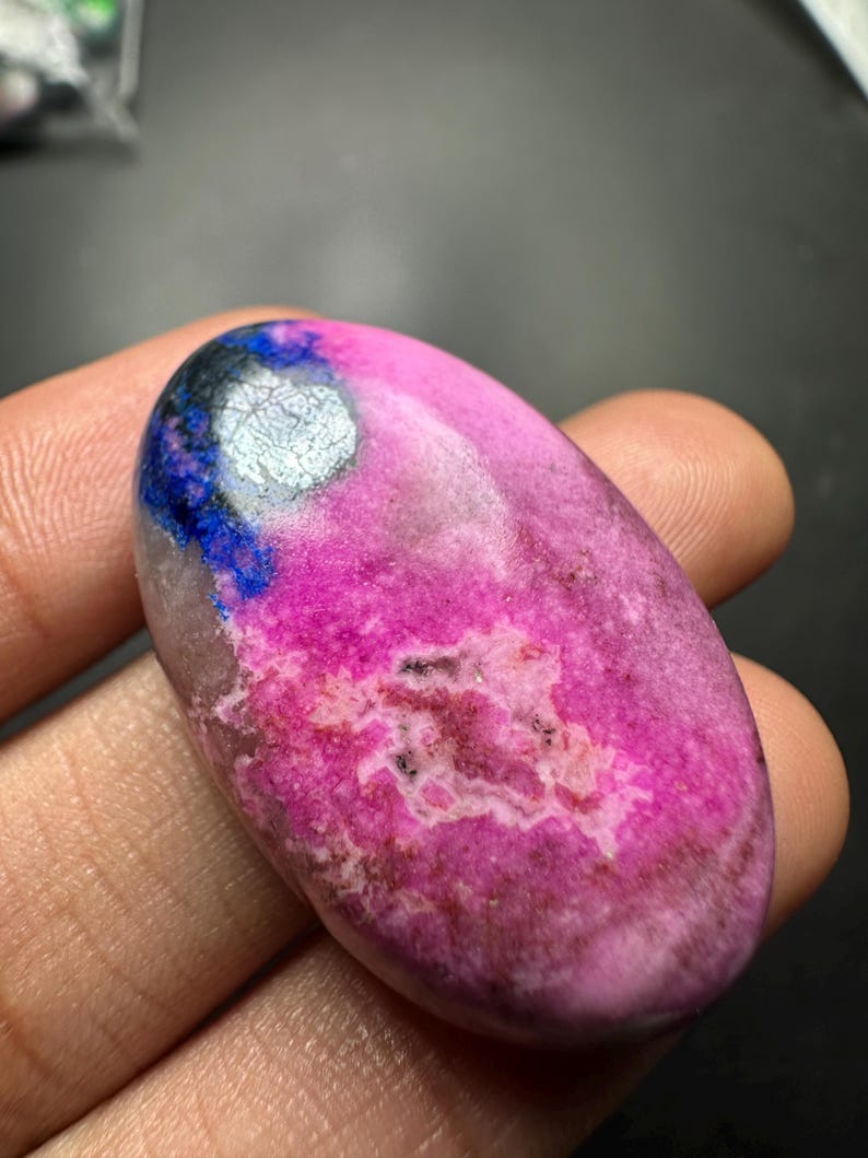 May include: An oval-shaped polished gemstone featuring a vibrant hot pink base color with mottled white and brown inclusions. A distinct patch of deep blue and metallic silver-grey mineral deposits is visible on the upper left side of the stone.