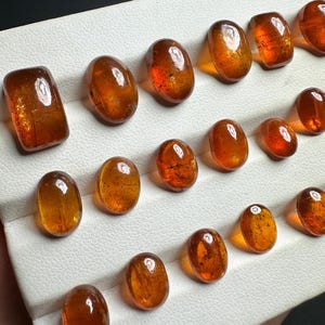 May include: A collection of 16 oval-shaped, translucent, orange-brown gemstones displayed on a white surface.