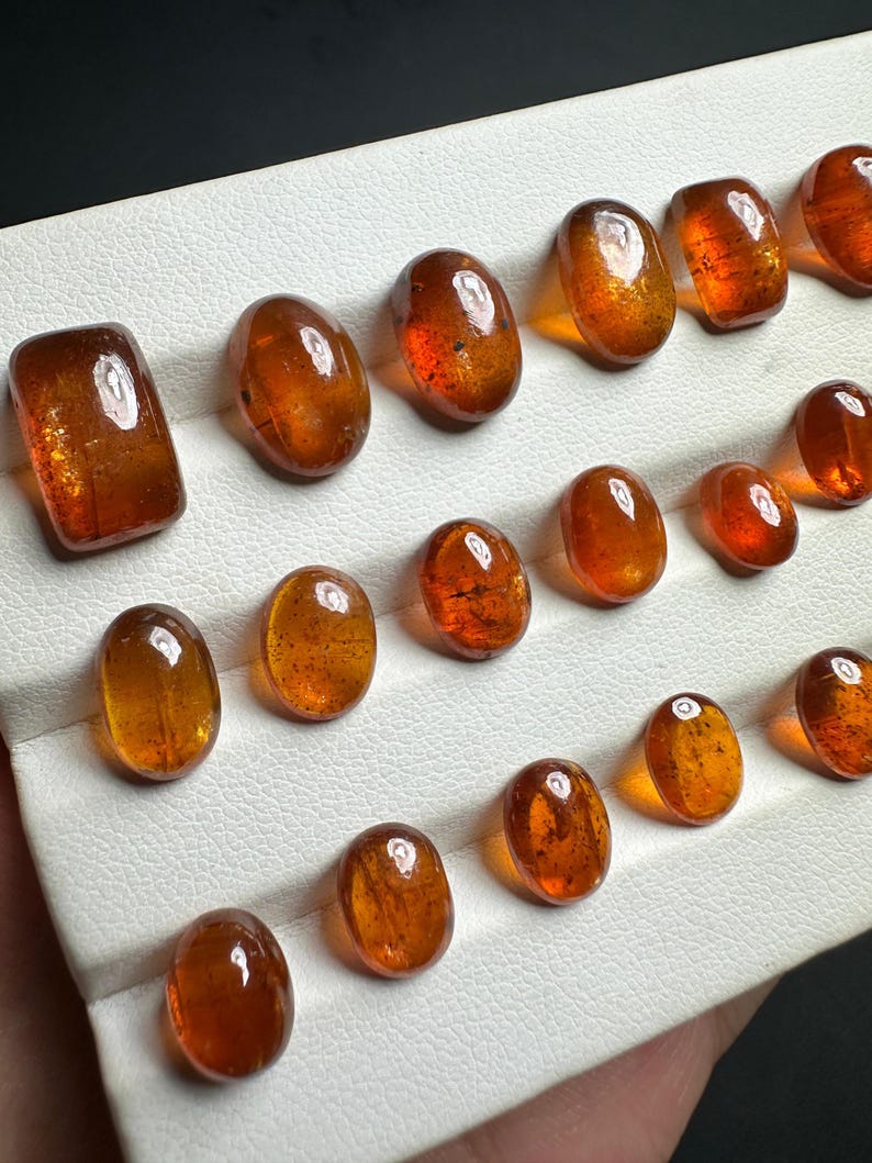 May include: A collection of 16 oval-shaped, translucent, orange-brown gemstones displayed on a white surface.