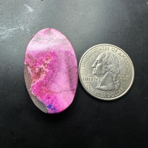 May include: An oval-shaped, polished pink stone cabochon with white and grey mineral inclusions, positioned next to a United States quarter coin for scale. The word Back is printed in black text in the upper left corner of the image.