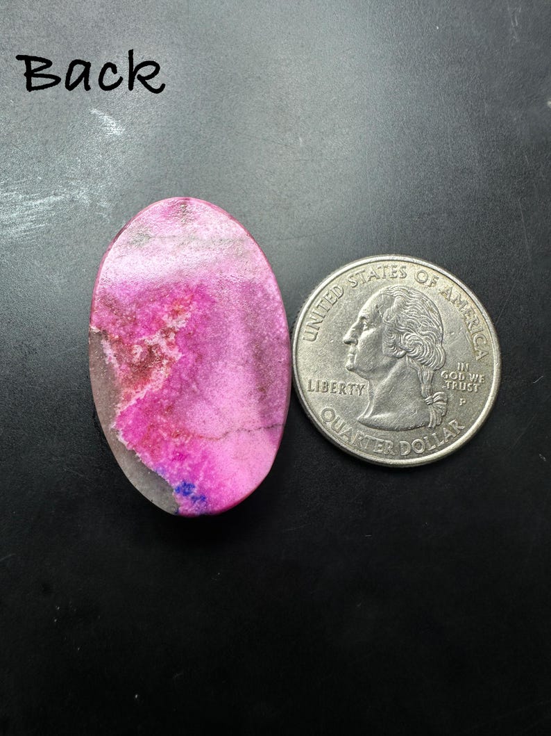 May include: An oval-shaped, polished pink stone cabochon with white and grey mineral inclusions, positioned next to a United States quarter coin for scale. The word Back is printed in black text in the upper left corner of the image.