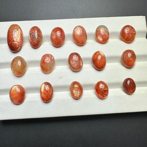 May include: A collection of 15 oval-shaped sunstone gemstones, ranging in color from orange to red, displayed on a white background.
