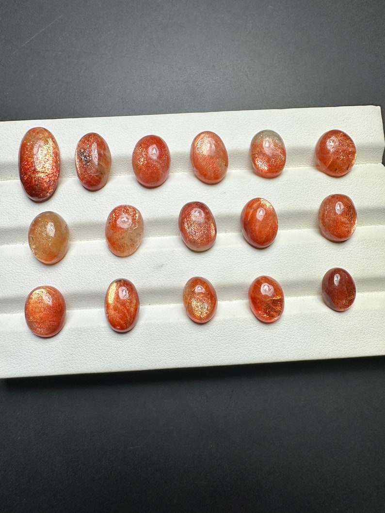 May include: A collection of 15 oval-shaped sunstone gemstones, ranging in color from orange to red, displayed on a white background.