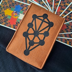 Puede incluir: Una funda naranja para cartas del tarot con un diseño geométrico negro. La funda rectangular está hecha de tela texturizada. Un mazo de cartas del tarot con ilustraciones coloridas es visible en el fondo. La funda está diseñada para guardar cartas del tarot.