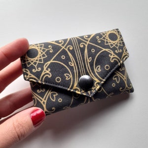 May include: A small, black fabric wallet with a gold geometric pattern and a black snap closure. The wallet is held in a hand with red nail polish. The design features swirling lines and circular shapes.