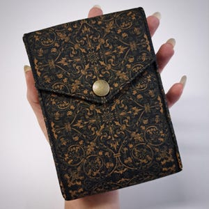 Puede incluir: Una cartera negra y dorada estampada con solapa y cierre de botón de latón. La cartera presenta un diseño ornamentado de estilo vintage. La cartera rectangular se sostiene en una mano.