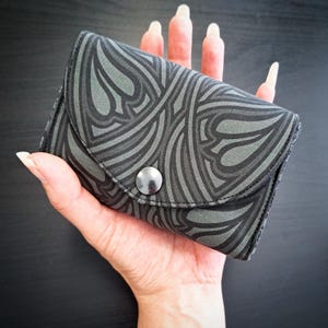 Puede incluir: Una pequeña cartera gris con un patrón abstracto repetido en gris más oscuro. La cartera tiene un cierre de botón a presión y se sostiene en una mano. El diseño presenta líneas y formas curvas, creando un aspecto artístico y fluido. Es un accesorio compacto.