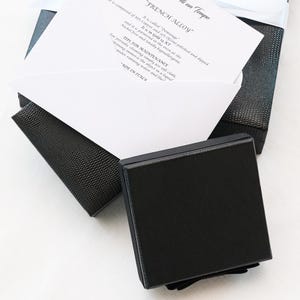 May include: A black square box with a textured surface, partially open, revealing a white card with text. The card details the composition and care instructions for a "FRENCH ALLOY" product, including "Demionge" and "Gold 14 KT".