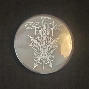 Op de afbeelding: Een ronde, grijze knoop met de bandnaam "Celtic Frost" in witte, gestileerde letters. De knoop heeft een wit, ingewikkeld geometrisch ontwerp in het midden, met scherpe, hoekige vormen. De knoop is waarschijnlijk een verzamelobject of accessoire.