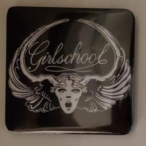 Puede incluir: Botón cuadrado negro con un gráfico blanco de una cara con la boca abierta y alas, con la palabra "Girlschool" en cursiva. El diseño es una ilustración estilizada de inspiración vintage.