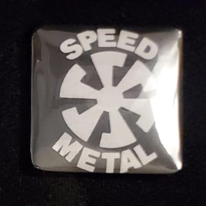 May include: A square, silver-coloured button featuring the words "SPEED METAL" in white, curved at the top and bottom. A white, stylised, eight-pointed starburst design is in the centre.