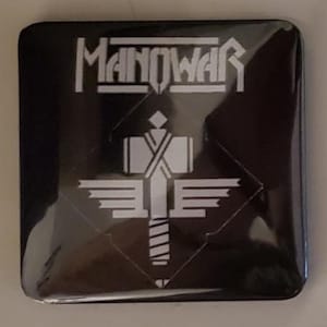 Könnte beinhalten: Schwarzer quadratischer Knopf mit dem Wort "MANOWAR" in weißen Blockbuchstaben oben. Darunter befindet sich eine weiße Grafik einer Person mit Flügeln und einem Schwert. Der Knopf ist wahrscheinlich ein Sammlerstück oder Fanartikel.