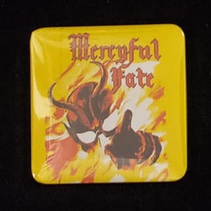 May include: Square yellow button with the text "Mercyful Fate" in a gothic font. The button features a red and black illustration of a horned demon in flames. The button has a glossy finish.