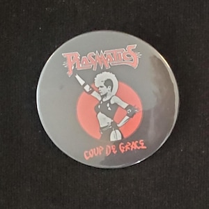 Könnte beinhalten: Ein runder, grauer Button mit dem Bandnamen "Plasmatics" in roter, stilisierter Schrift. Der Button hat einen roten Kreis mit einer Grafik einer Frau in einer Punkrock-Pose. Der Text "COUP DE GRACE" ist unten in Rot geschrieben.