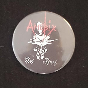 May include: A round, gray button with the word "AMIBIX" in red, stylized text. Below the text is a white illustration of a face with a crown. The words "NO GODS" and "NO MASTERS" are printed in white.