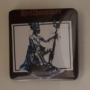 May include: Square button with a dark brown border featuring the band name "Hellhammer" in gothic font. The center shows a black and blue illustration of a skeletal figure holding a staff.