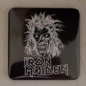 May include: Square black coaster featuring a white illustration of a skeletal face with wild hair. The words "IRON MAIDEN" are printed in white, stylized lettering below the image. The coaster has rounded corners.