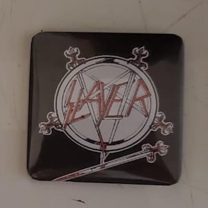 Könnte beinhalten: Quadratischer schwarzer Knopf mit dem Wort "SLAYER" in roter, stilisierter Schrift. Der Text befindet sich in einem weißen Pentagramm mit einem Schwert und dekorativen Elementen. Der Knopf hat abgerundete Ecken.