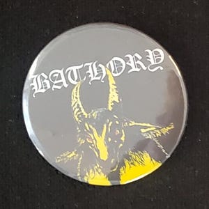 May include: A round, gray button with the word "BATHORY" in white gothic lettering. The button features a yellow and black illustration of a horned goat with red eyes.
