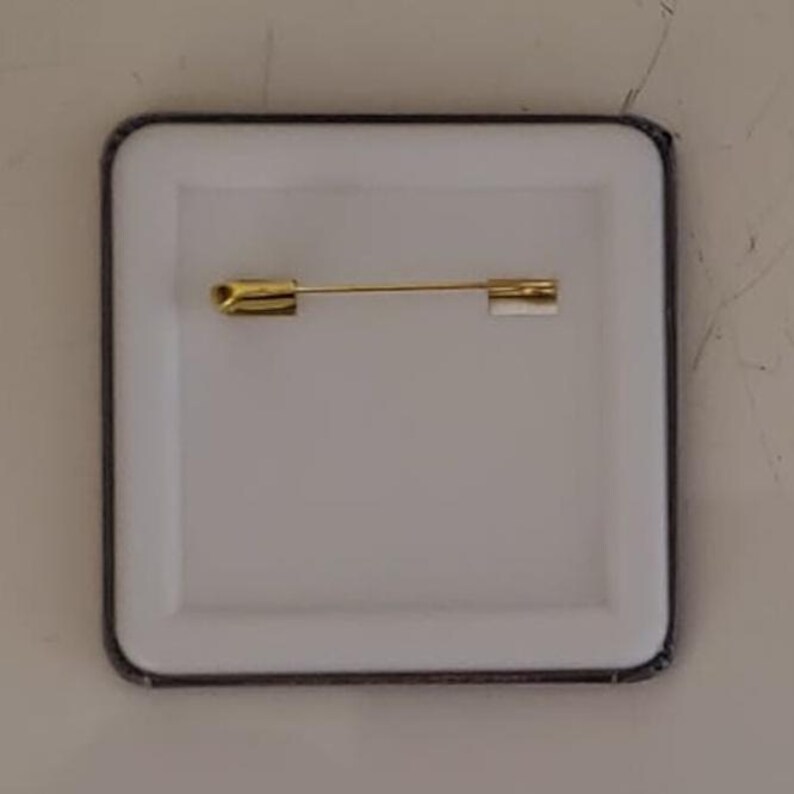 Puede incluir: Un bot&oacute;n cuadrado con un centro blanco y un alfiler dorado en la parte posterior. El bot&oacute;n tiene un borde negro y un dise&ntilde;o sencillo, adecuado para diversos usos.