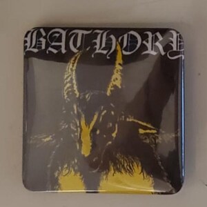 以下が含まれることがあります： 白いゴシック体の文字で「BATHORY」と書かれた正方形のボタン。角のあるヤギの黒と黄色のイラストが描かれています。背景は暗いです。