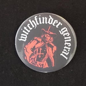 May include: A round, black button with the words "witchfinder general" in white around the edge. The button features a red illustration of a man in a hat and cloak. The button is likely a pin or badge.