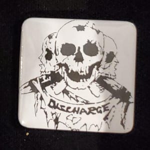 May include: Square, white coaster with a black graphic of three skulls. The word "DISCHARGE" is printed in black on the coaster. The coaster has rounded corners and a glossy finish.