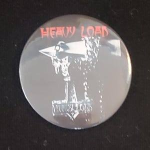 Peut inclure: Un bouton rond gris avec les mots "HEAVY LOAD" en rouge en haut. En dessous, une illustration blanche représente un marteau et une enclume. Les mots "PULLY LOAD" sont en blanc en bas.