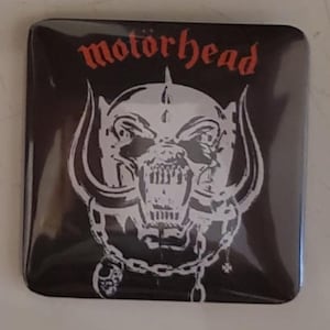 Puede incluir: Botón cuadrado negro con el nombre de la banda "motorhead" en fuente gótica roja sobre una calavera blanca con colmillos y una cadena. El diseño de la calavera es un símbolo reconocible de la banda.