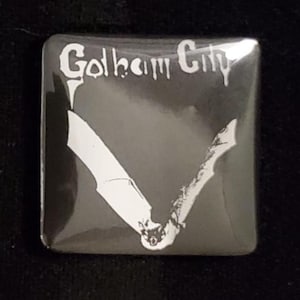 Puede incluir: Bot&oacute;n cuadrado con fondo negro e ilustraci&oacute;n de murci&eacute;lago blanco. El texto "Gotham City" est&aacute; escrito en blanco en la parte superior del bot&oacute;n. El bot&oacute;n es cuadrado con esquinas redondeadas.