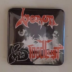May include: Square black button with the word "Venom" in red, stylized lettering at the top. Below, the word "Bloodlust" is in red, with a white and black graphic of three faces. The button has a glossy finish.