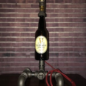May include: A unique industrial-style lamp made from a Guinness beer bottle, metal pipes, and a blue valve. The lamp has a red power cord and a brown plug. The bottle features a gold and black label.